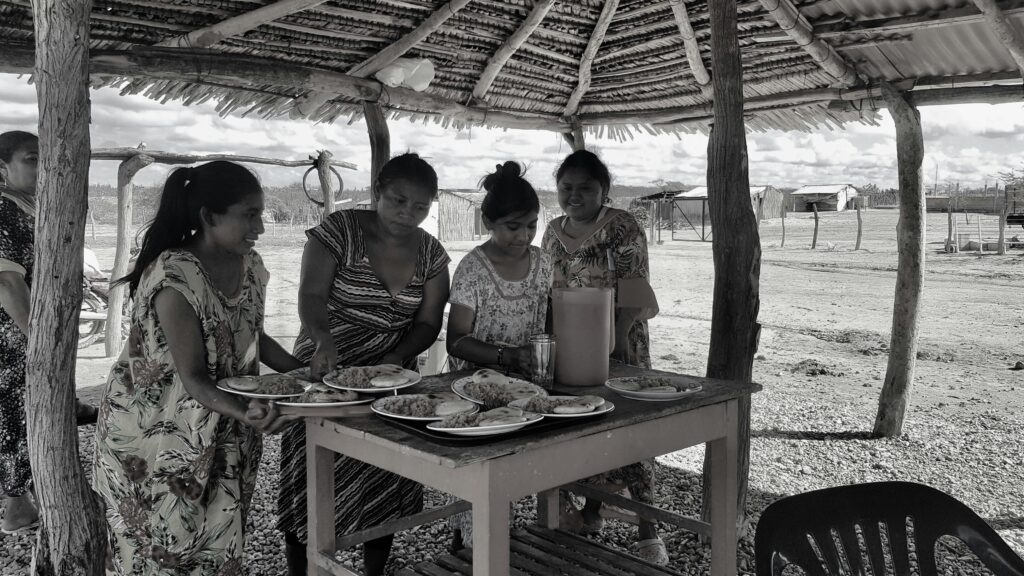 Four women serve traditional food under a rustic outdoor shelter.