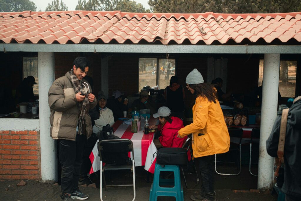 People enjoying a casual outdoor gathering in a rustic cafe setting under a red-tiled shelter.